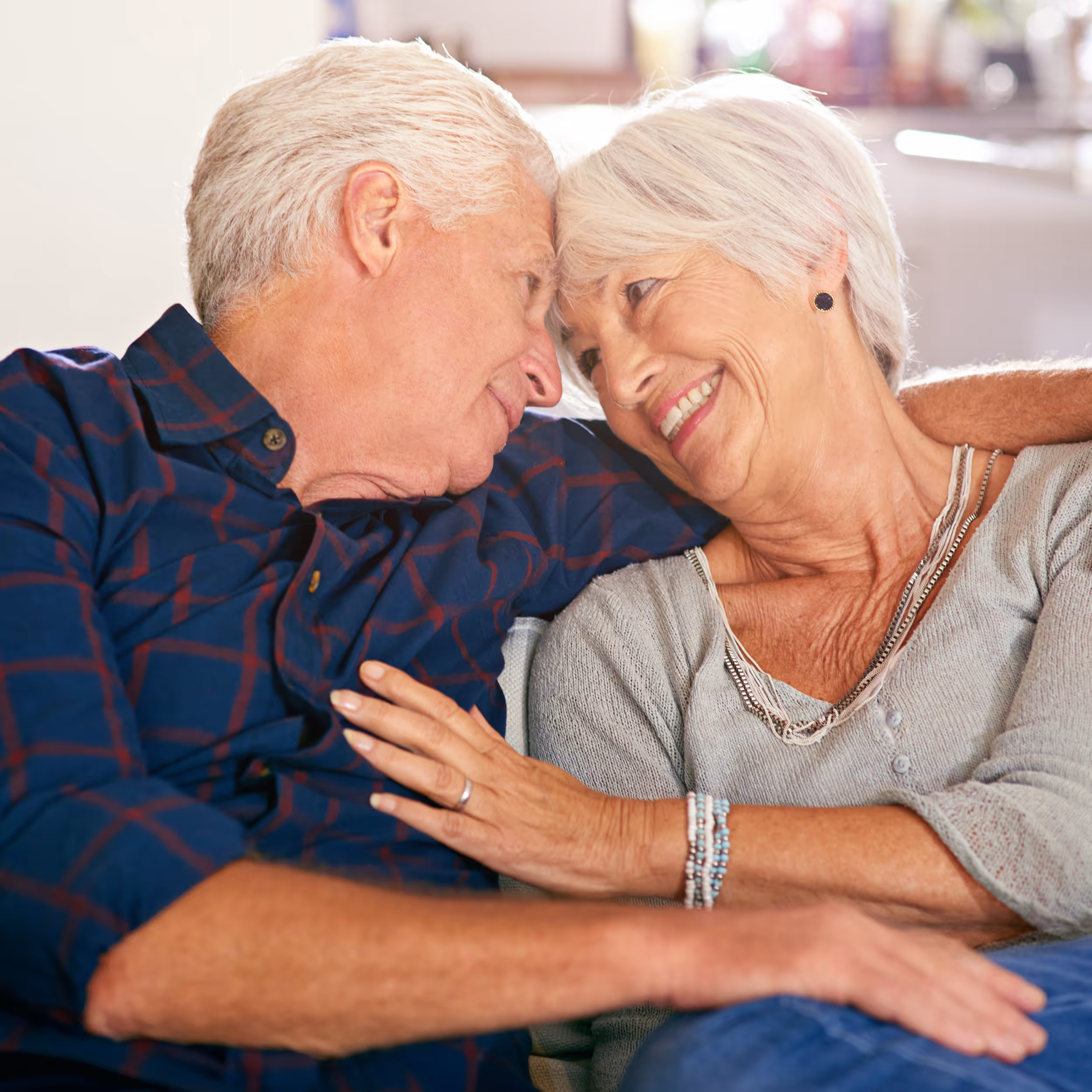 Elderly couple sitting close together on a couch, smiling and touching foreheads in an affectionate embrace.