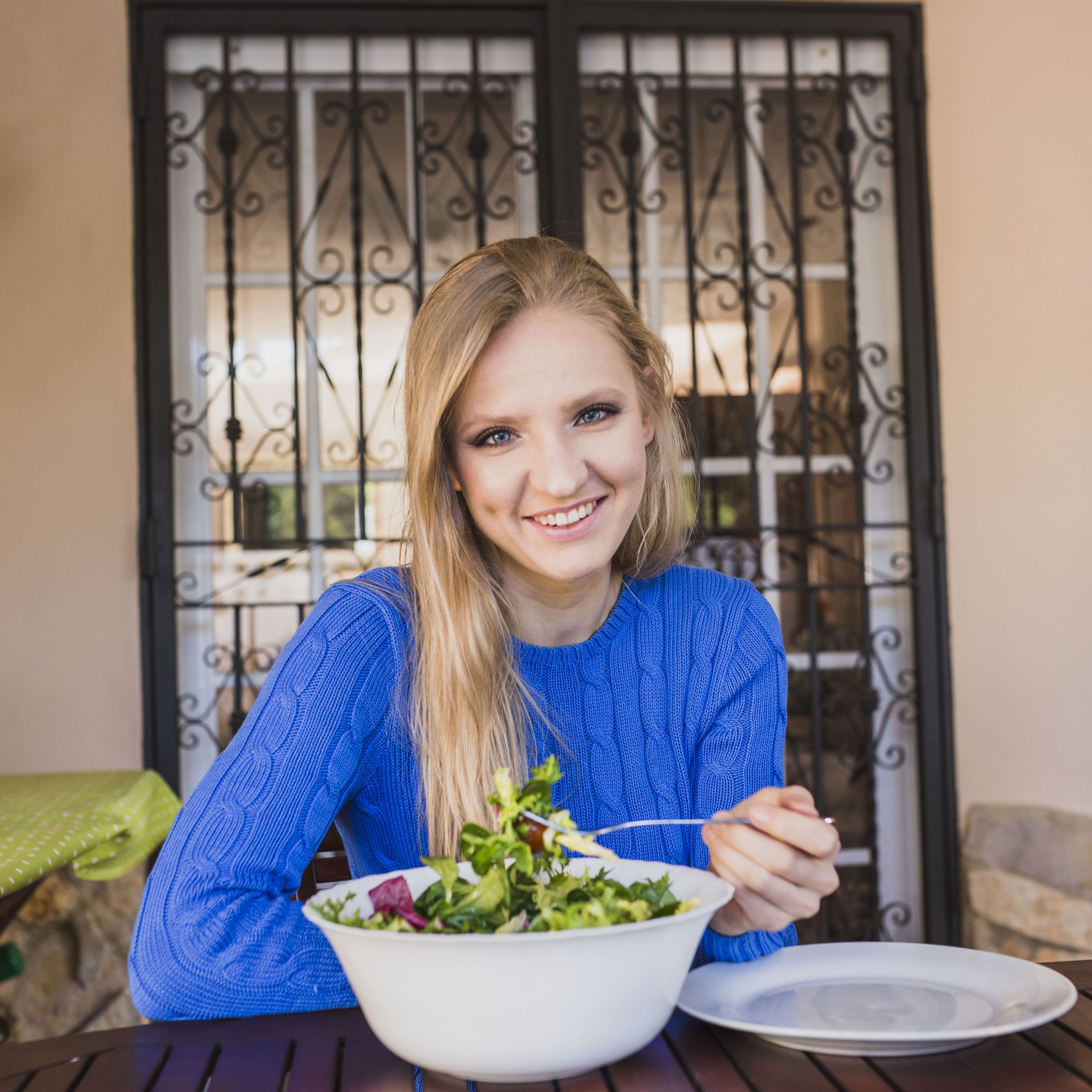 Smiling woman in blue sweater sitting at a table outdoors with a large bowl of fresh salad.