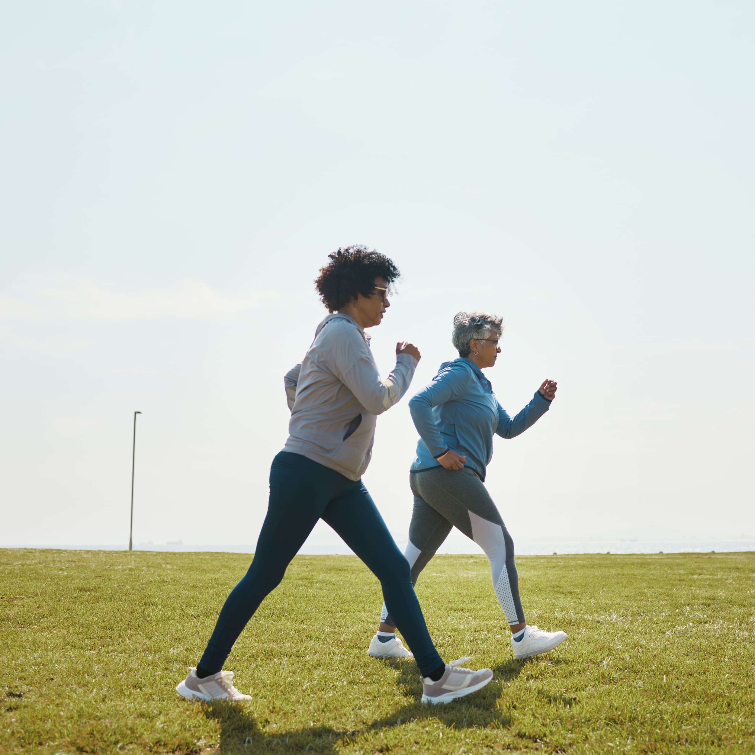Two women power walking on grassy field under clear sky, wearing athletic wear and sneakers.