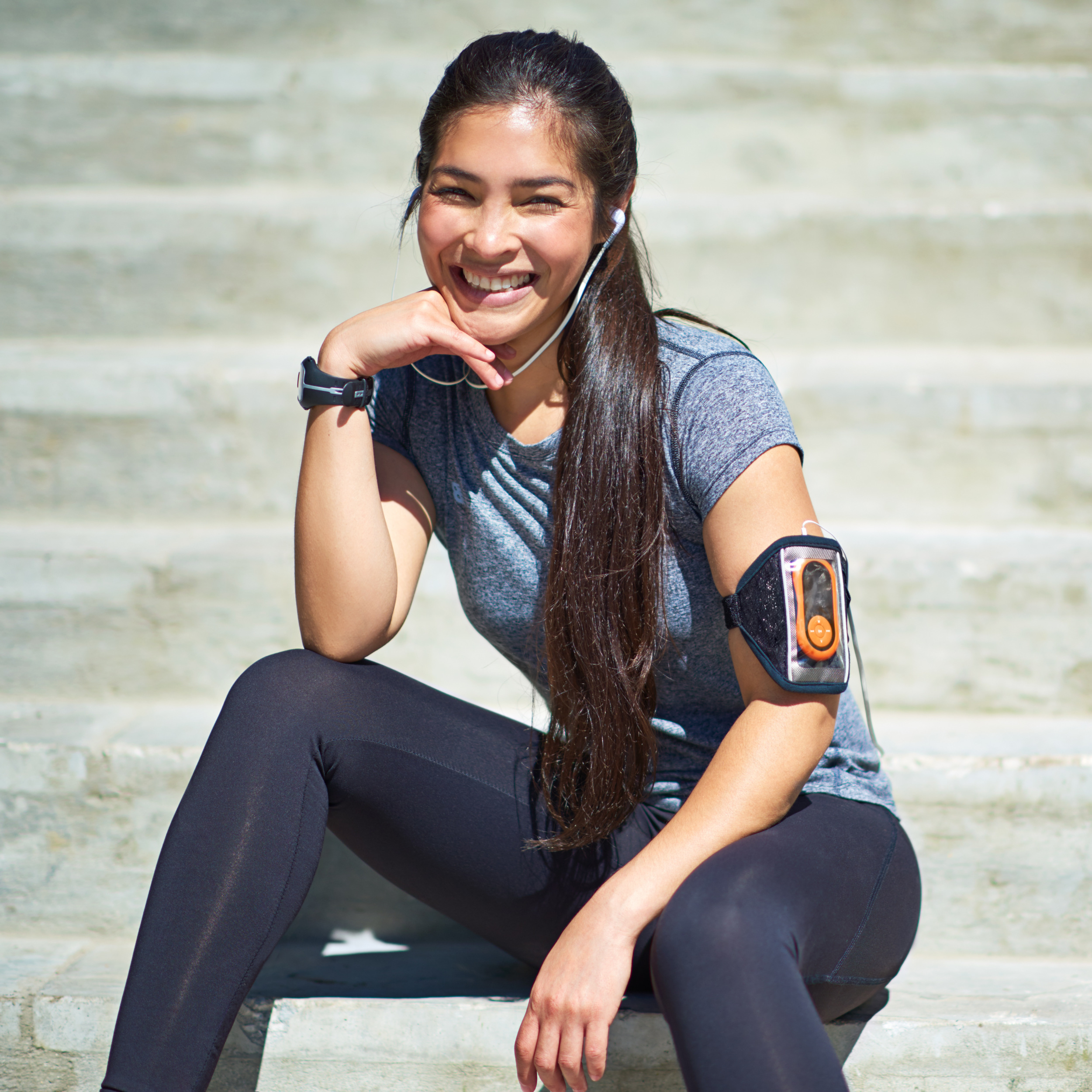 Smiling woman in workout clothes sitting on outdoor steps with earphones and fitness tracker.