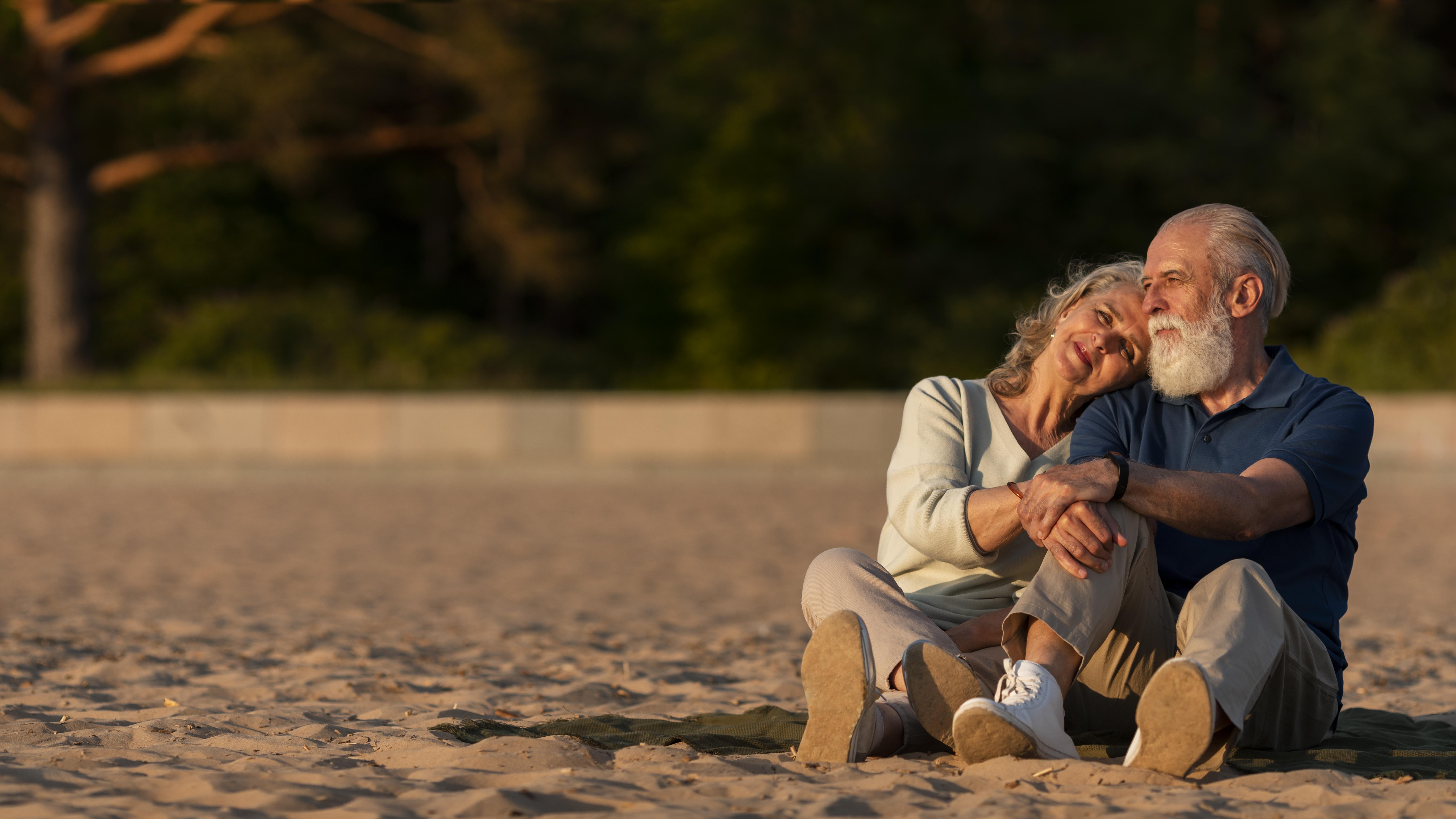 Elderly couple sitting closely on sand with the woman resting her head on the man's shoulder at sunset.