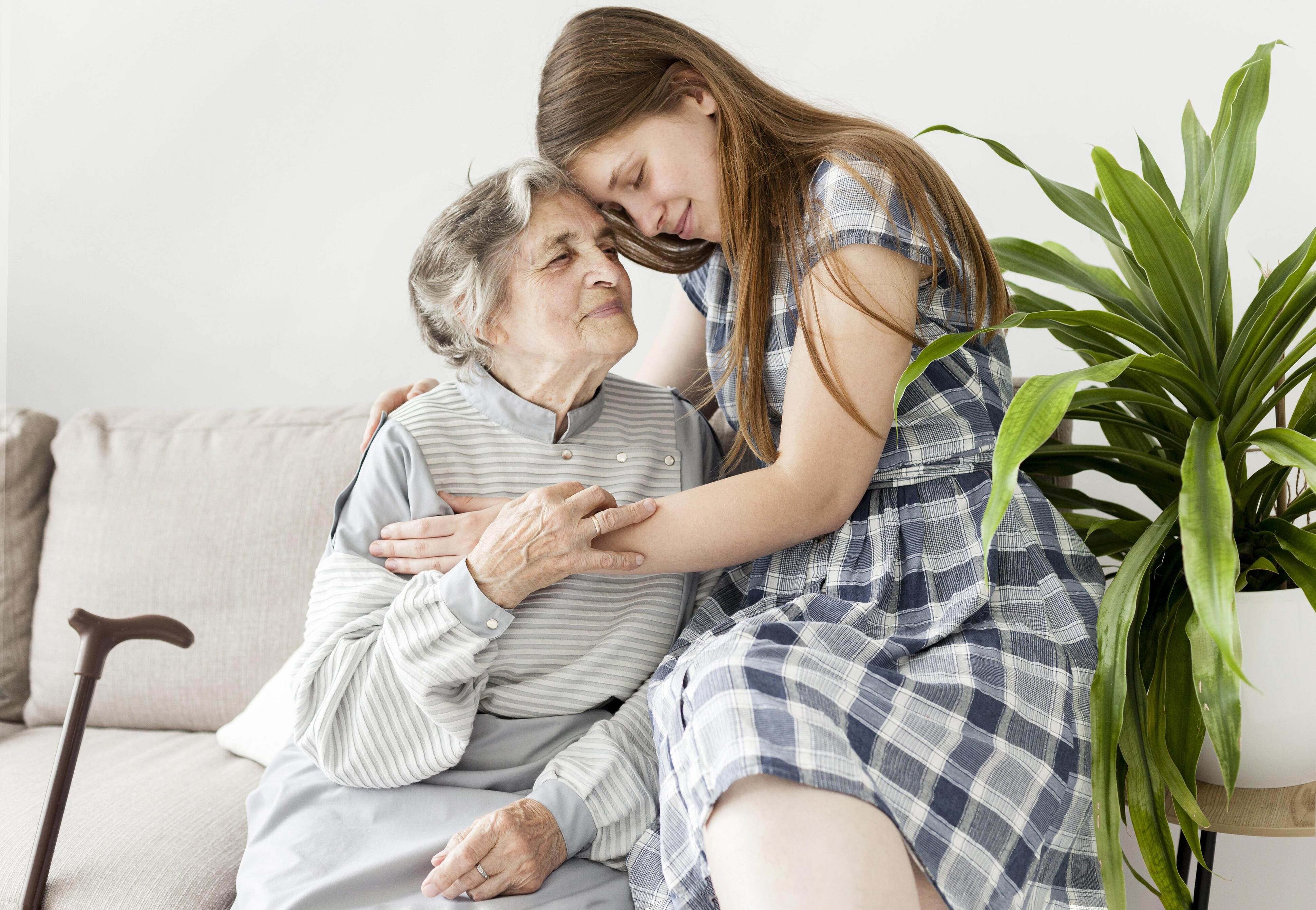 Elderly woman sitting on a couch warmly embracing a young girl in a plaid dress, with a walking cane resting beside the couch and a green potted plant nearby.
