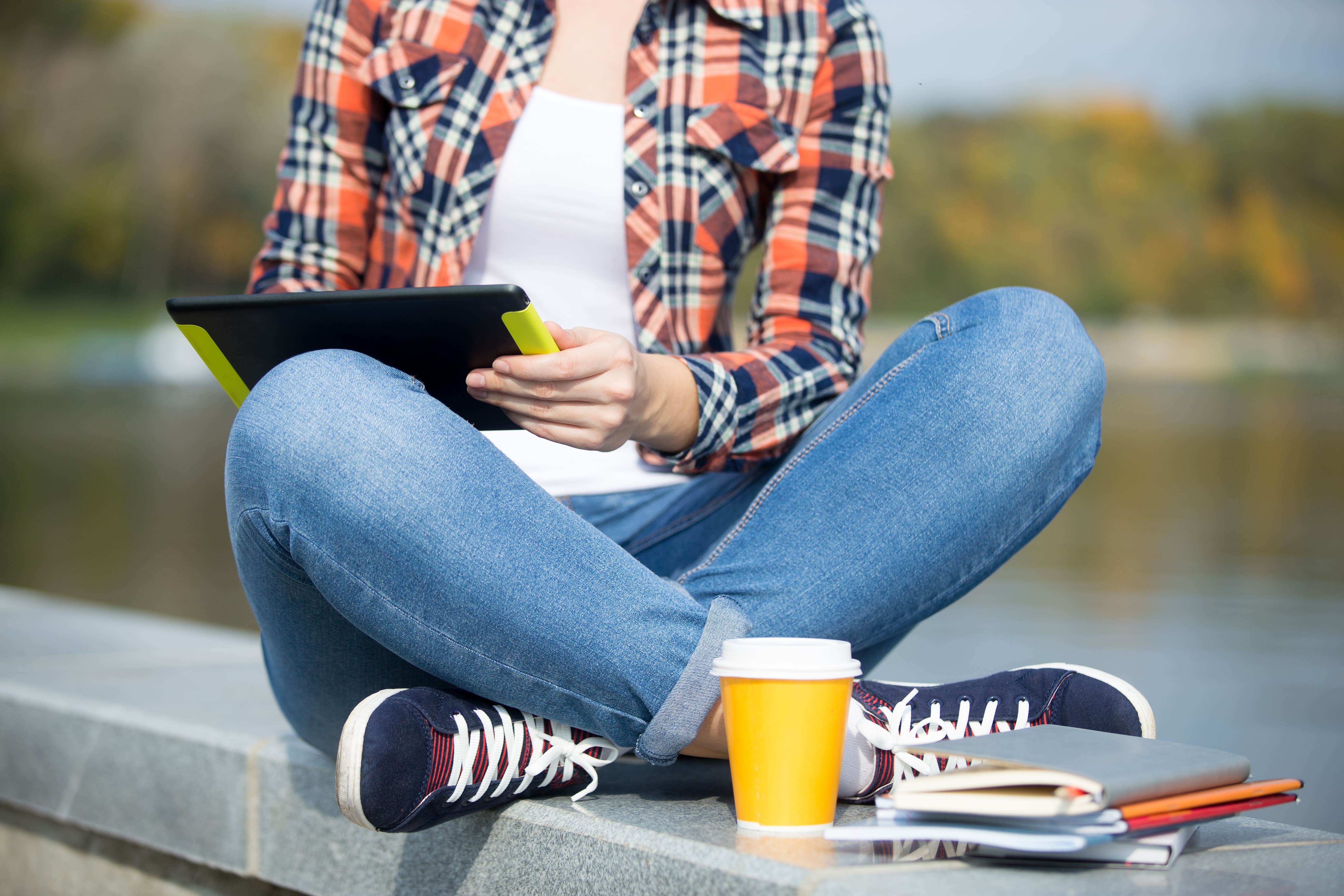 Person sitting cross-legged on a stone ledge by water, holding a tablet, with a yellow coffee cup and notebooks beside them.