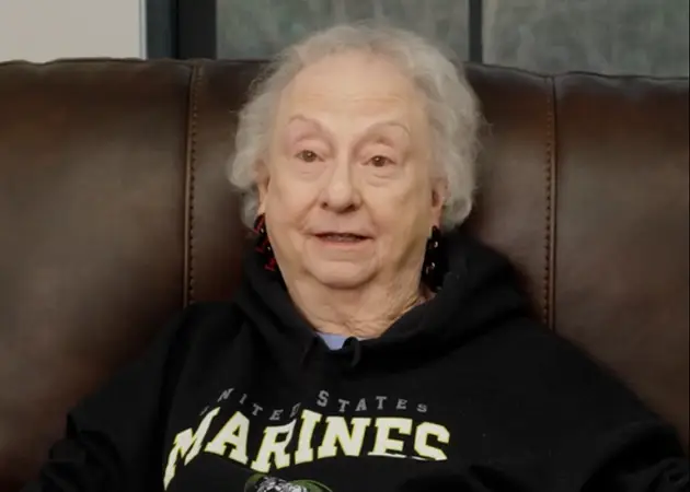 Elderly woman with curly white hair wearing a black United States Marines hoodie, sitting on a brown leather chair.