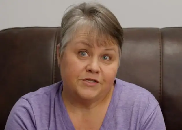 Middle-aged woman with short gray hair wearing a purple shirt sitting on a brown leather chair.