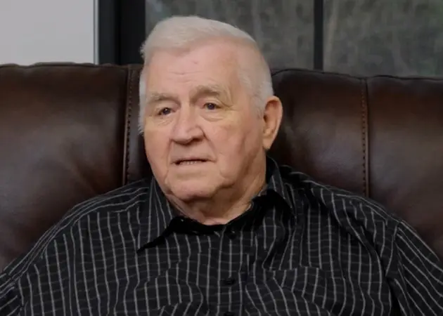 Elderly man with white hair wearing a black striped shirt sitting on a brown leather couch.