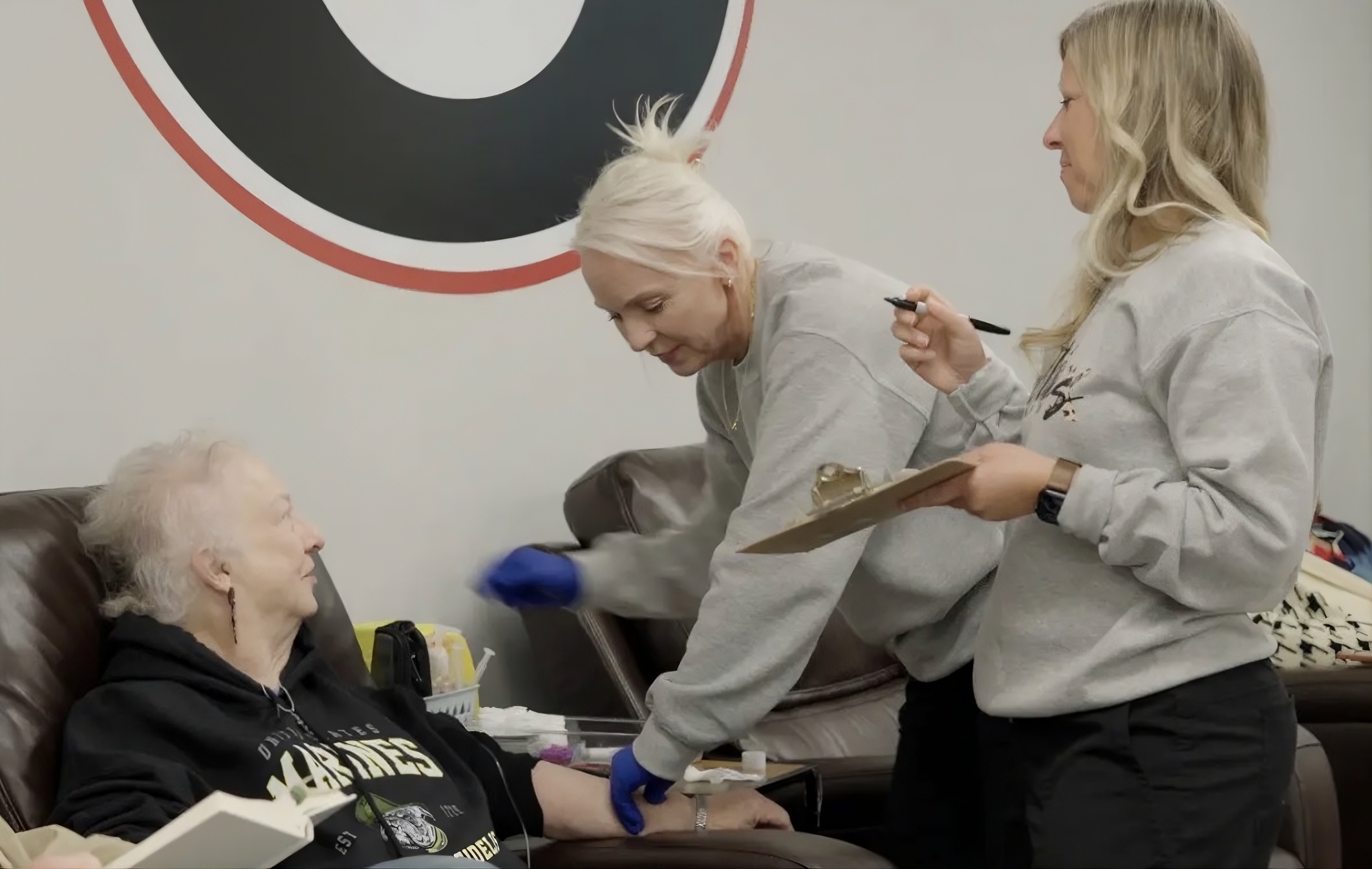 Two healthcare workers attending to an elderly woman seated on a leather chair, one preparing her arm while the other holds a clipboard and pen.
