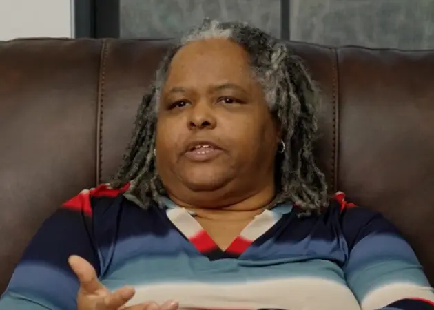 Person with gray and black dreadlocks and a striped blue, red, and white shirt speaking while seated on a brown couch.
