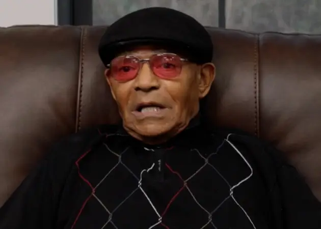 Elderly man wearing a black hat and red-tinted glasses sitting on a brown leather chair.