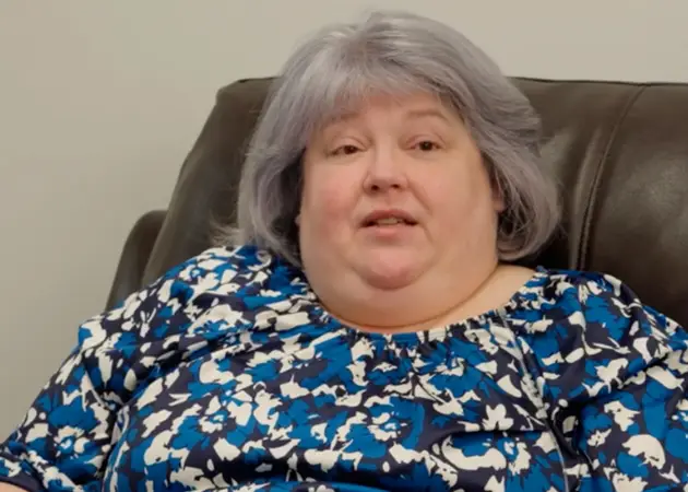 Middle-aged woman with gray hair wearing a blue and white floral blouse sitting on a brown chair against a beige background.