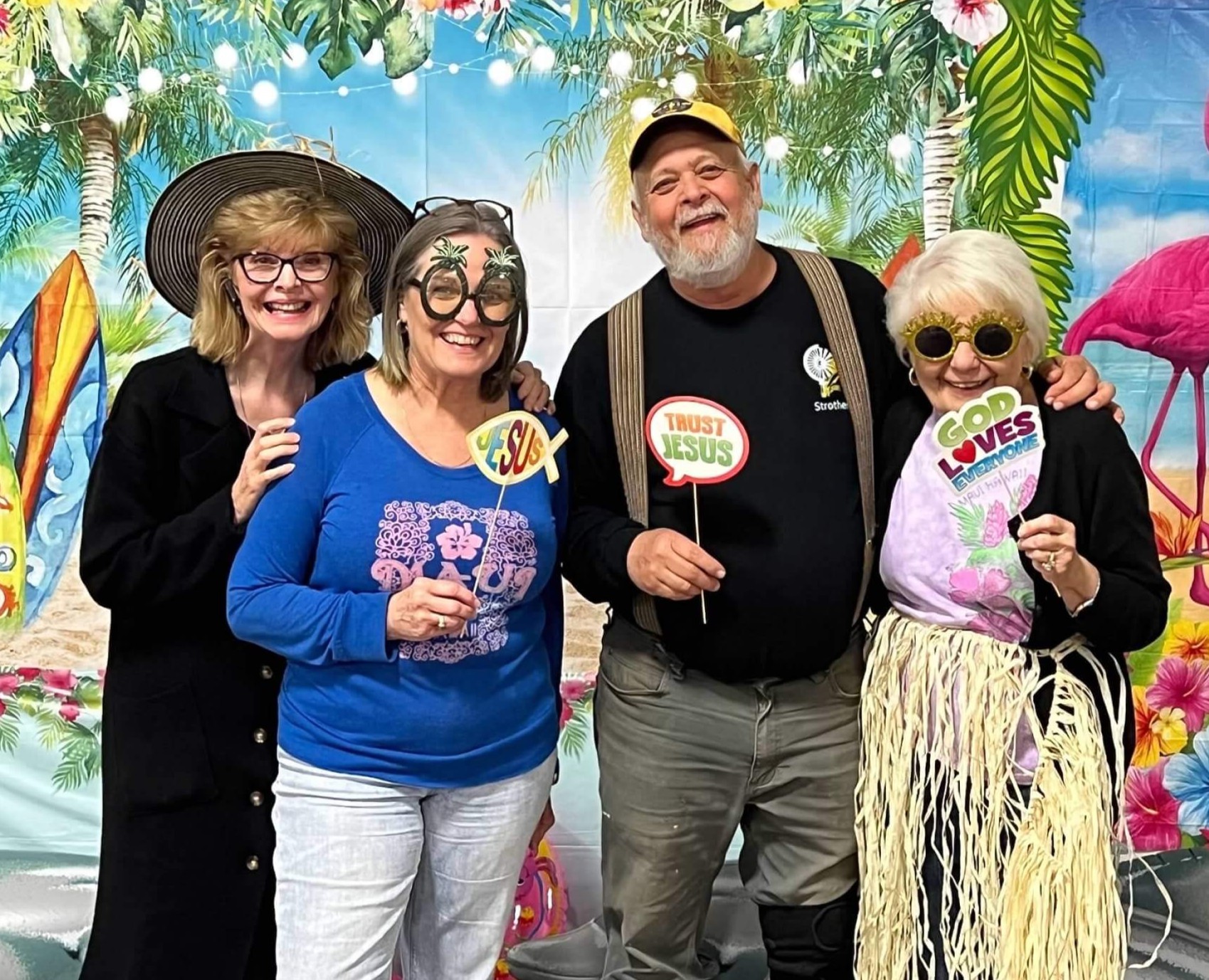 A group of four members of Redeemer Lutheran Church in Arkansas City, Kansas, holding props that say "Jesus" "God Loves everyone" and "Trust Jesus". They're also wearing sill, large, fake glasses and posing in front of a tropical backdrop. 