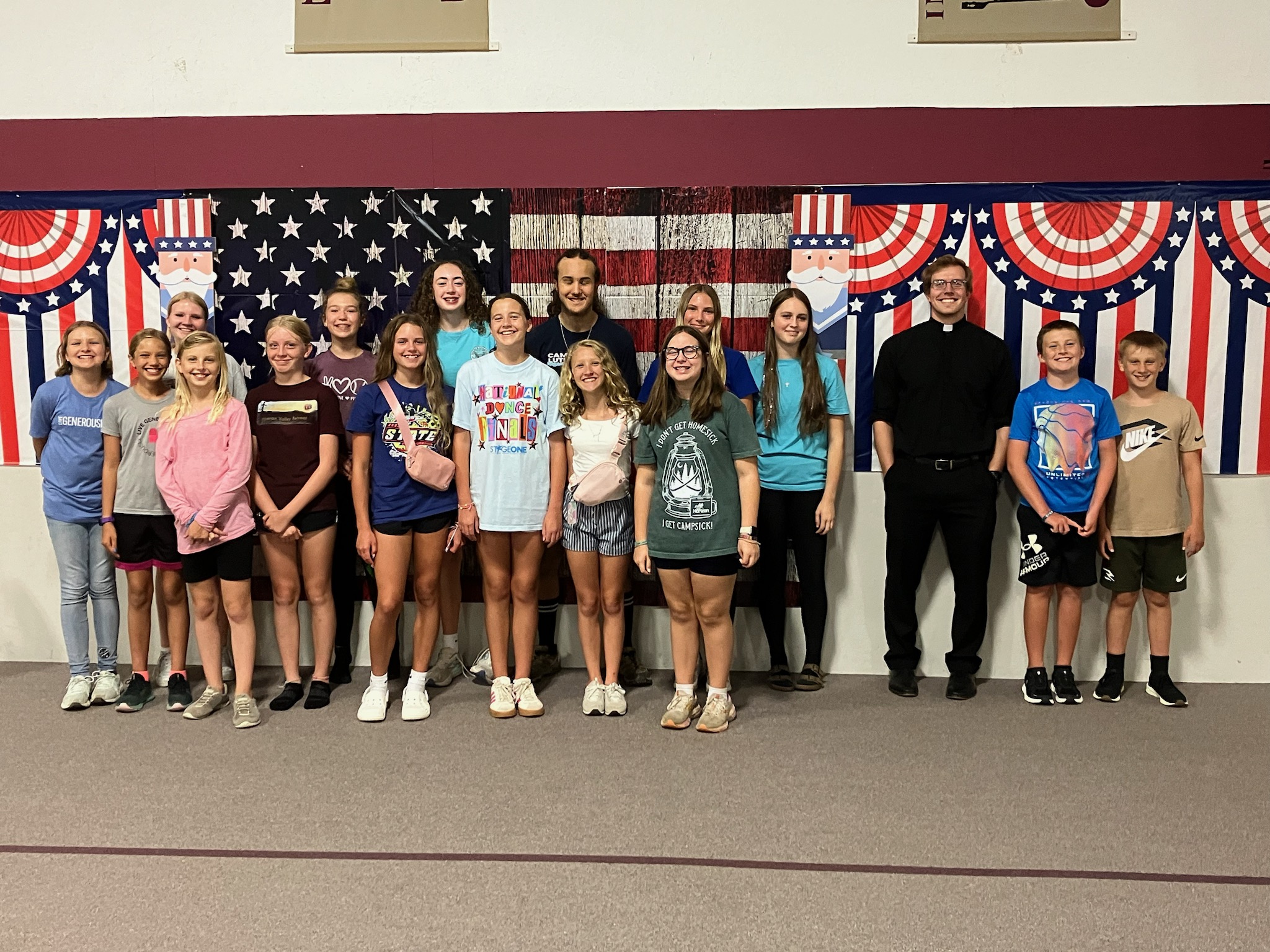 The youth group of Redeemer Lutheran Church, Arkansas City, Kansas and Pastor Warneke stand in front of an American flag in their gym. 