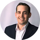 Smiling man wearing a dark suit jacket and white shirt against a light background.