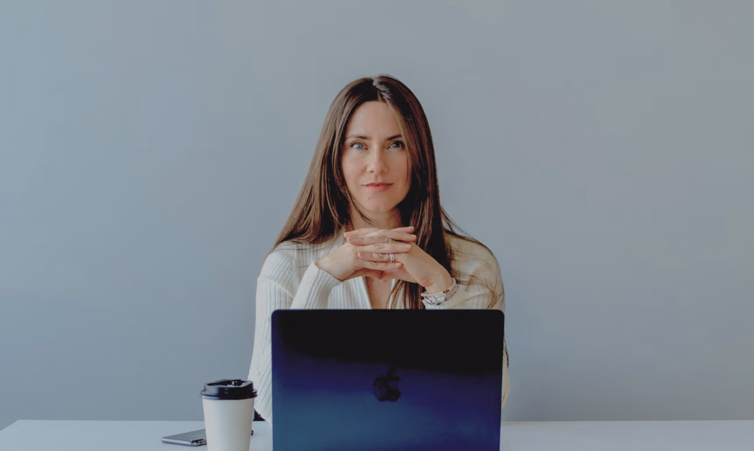 Woman with long brown hair sitting behind a laptop, with a coffee cup and smartphone on the table.