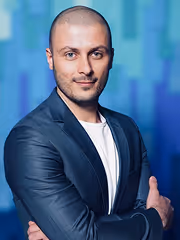 Confident man with a shaved head wearing a dark blazer and white shirt, standing with arms crossed against a blue blurred background.