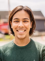 Young person with medium-length dark hair smiling outdoors, wearing a green sweater.