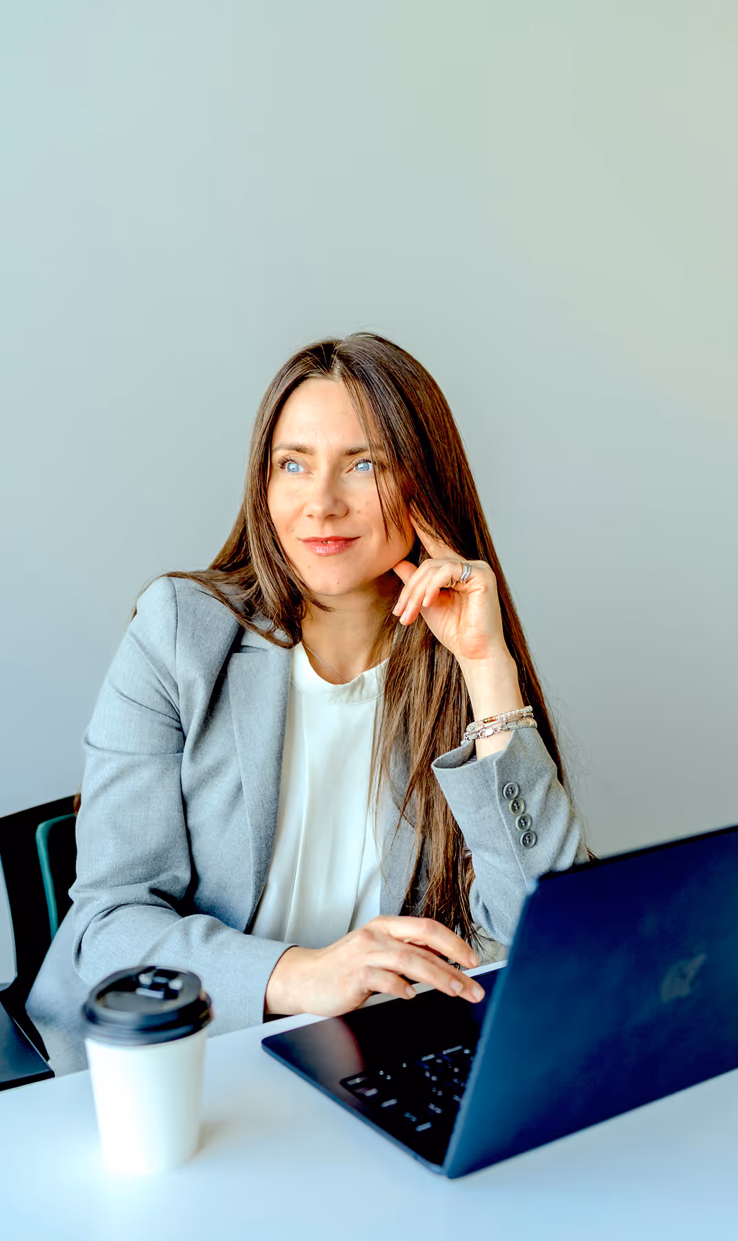 Woman in gray blazer sitting at a desk with a laptop and a takeaway coffee cup, looking thoughtfully to the side.