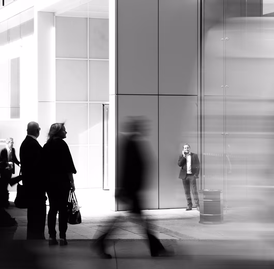 Black and white photo of a man standing and talking on a phone near a building wall with blurred people walking in the foreground.