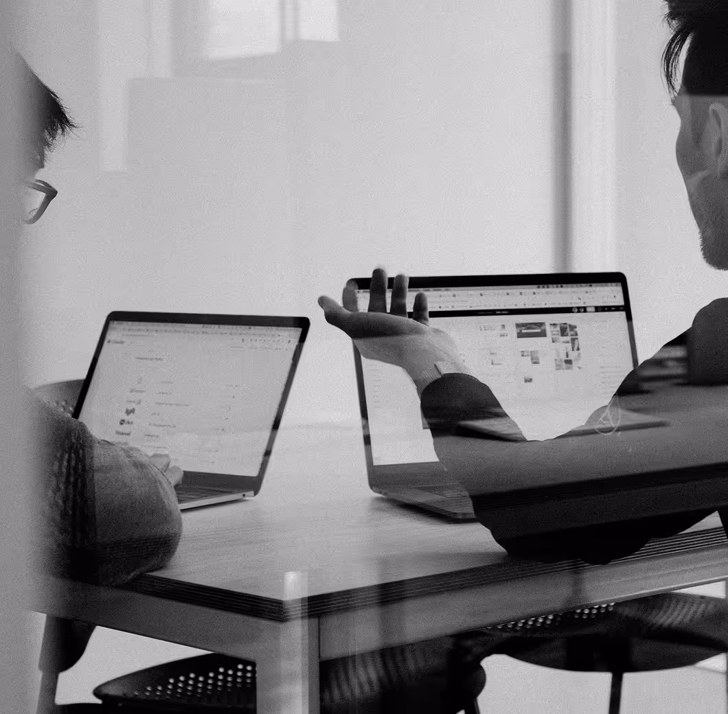 Two people working on laptops at a table, one gesturing with their hand during discussion.