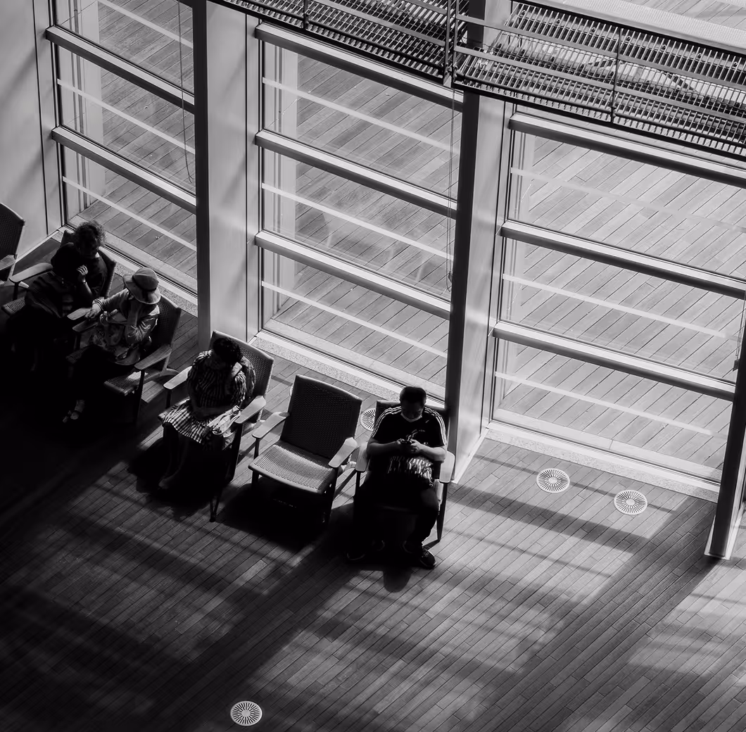 Black and white photo of four people sitting spaced apart on chairs by large floor-to-ceiling windows with sunlight casting shadows on the wooden floor.