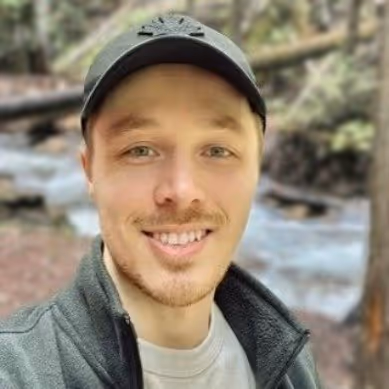 Young person with medium-length dark hair smiling outdoors, wearing a green sweater.