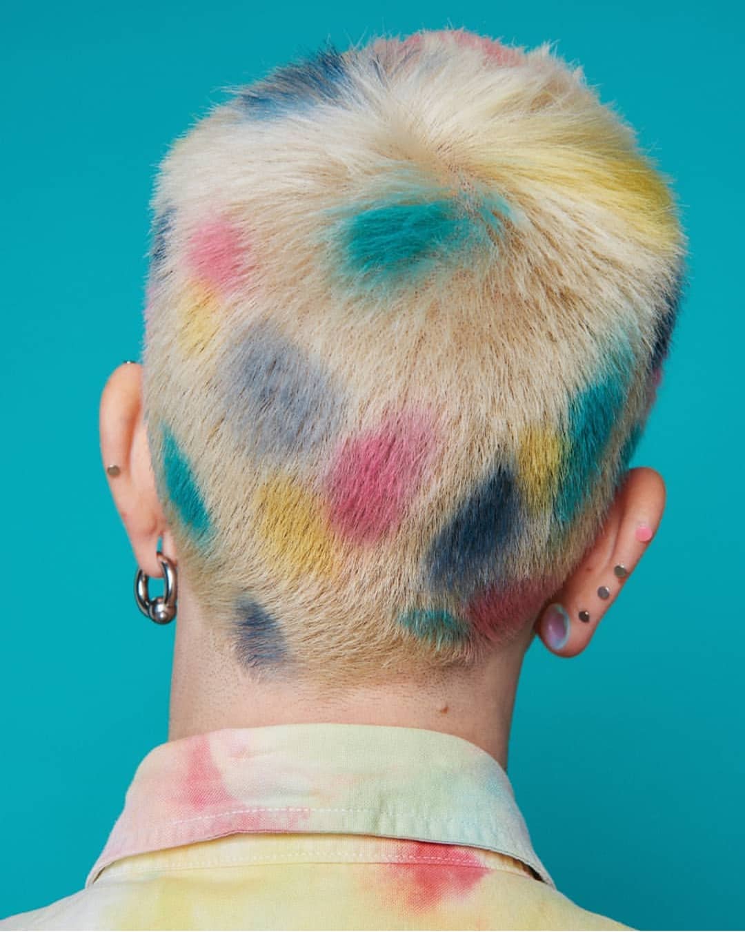 Back of a person's head with short blond hair dyed with multicolored patches including blue, pink, yellow, and gray against a teal background. Image by Panos Damaskinidis