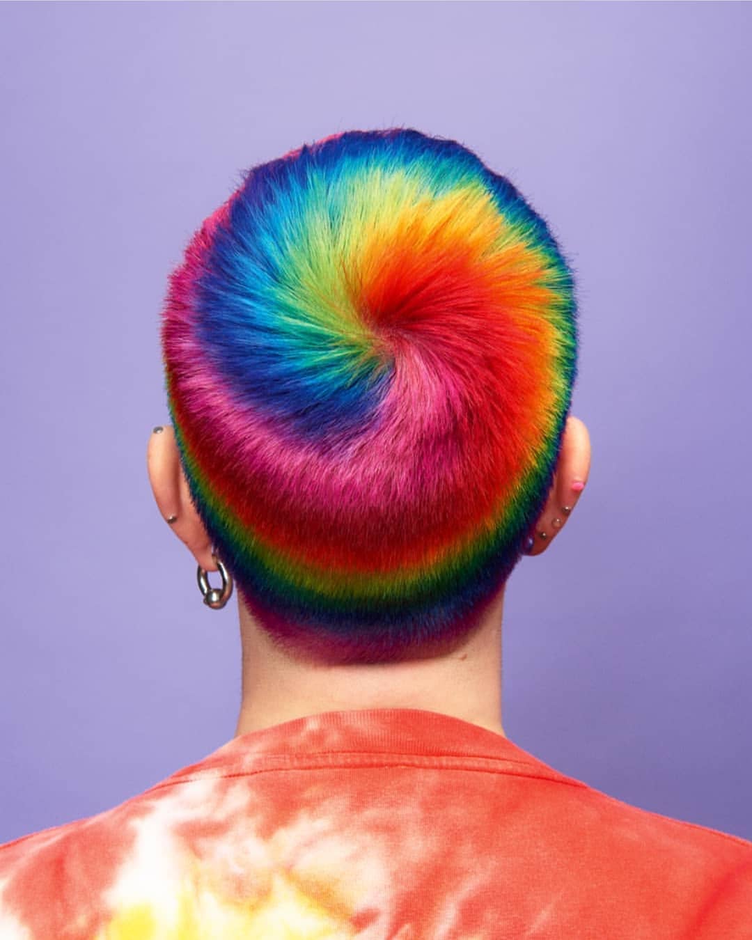 Back view of a person with short hair dyed in a rainbow spiral pattern, wearing an orange tie-dye shirt with purple background. Image by vPanos Damaskinidis
