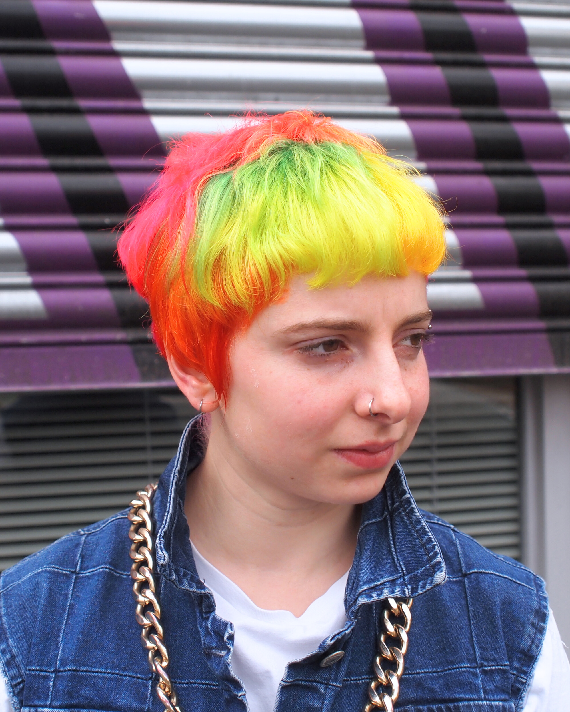 Person with short hair dyed in bright neon colours including pink, orange, yellow, and green, wearing a denim vest and a large gold chain necklace.