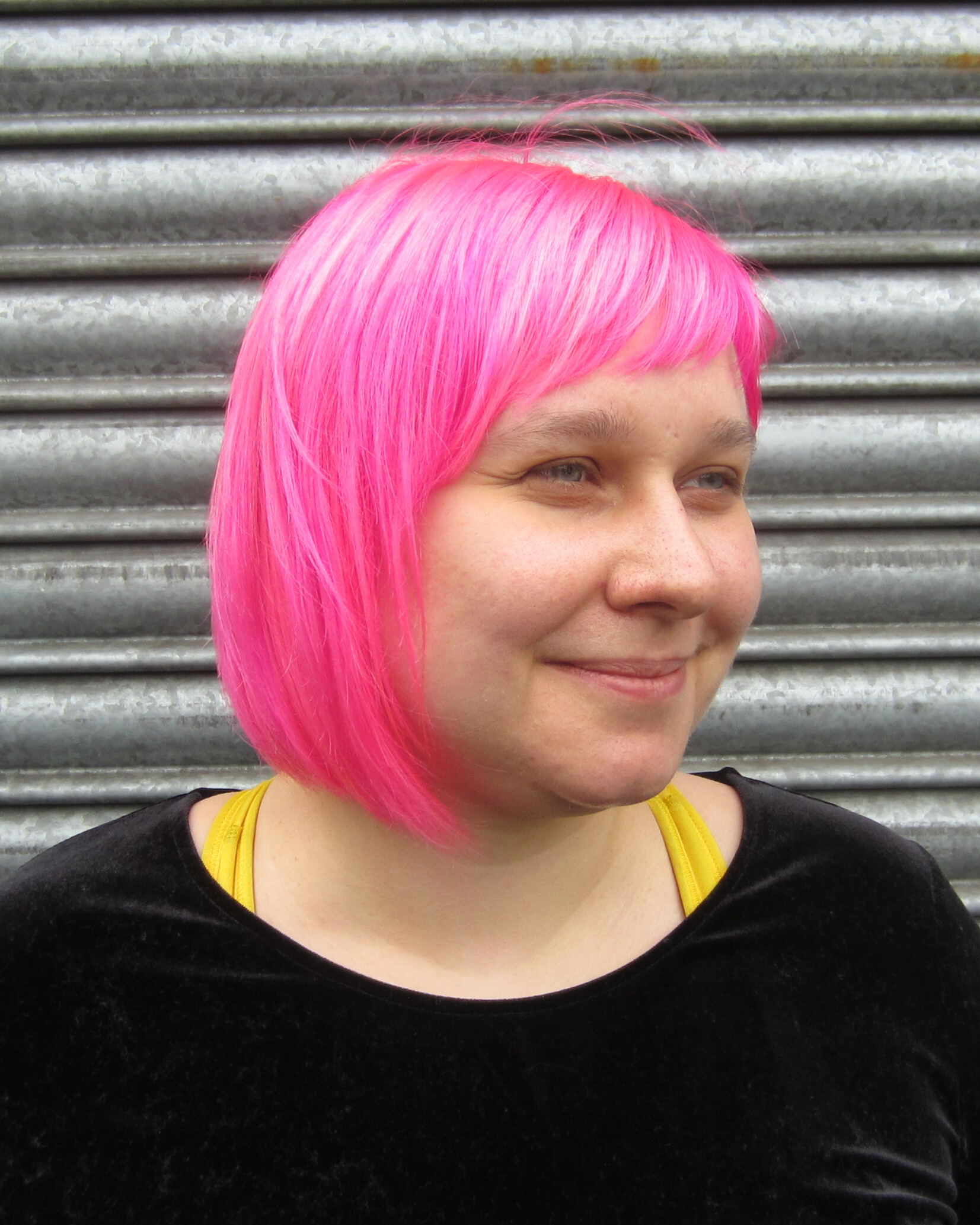 Person with bright pink bob haircut smiling, wearing black top standing in front of metal shutter.