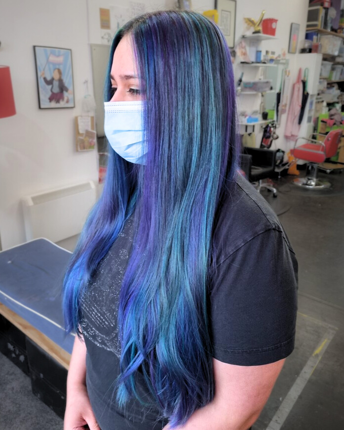 Woman with very long dark hair with highlights of different shades of blue, green and purple. Wearing a face mask and a black t-shirt in a hair salon.