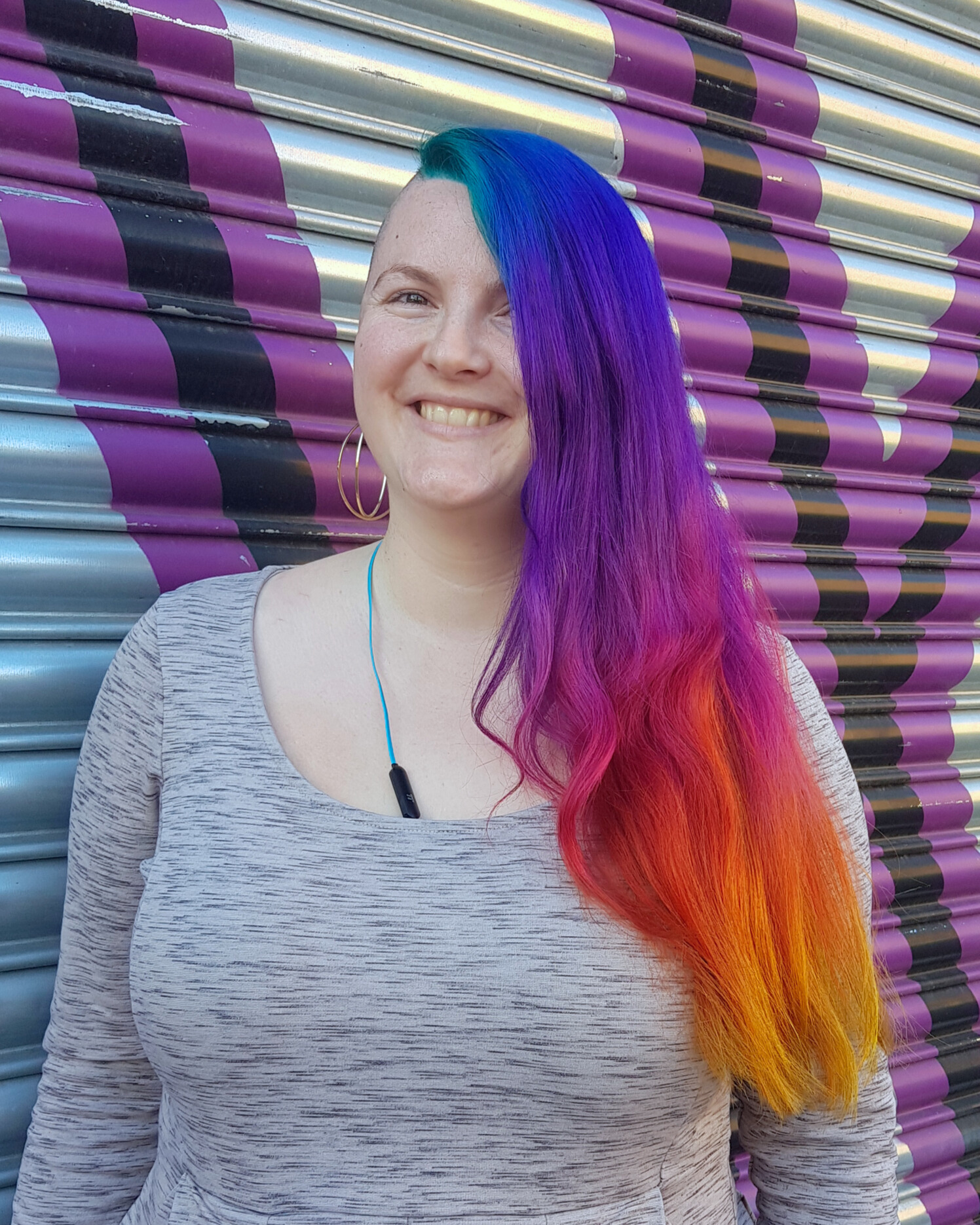 Smiling woman with long hair dyed blue, purple, pink, orange, and yellow standing in front of a purple and black striped metal shutter.
