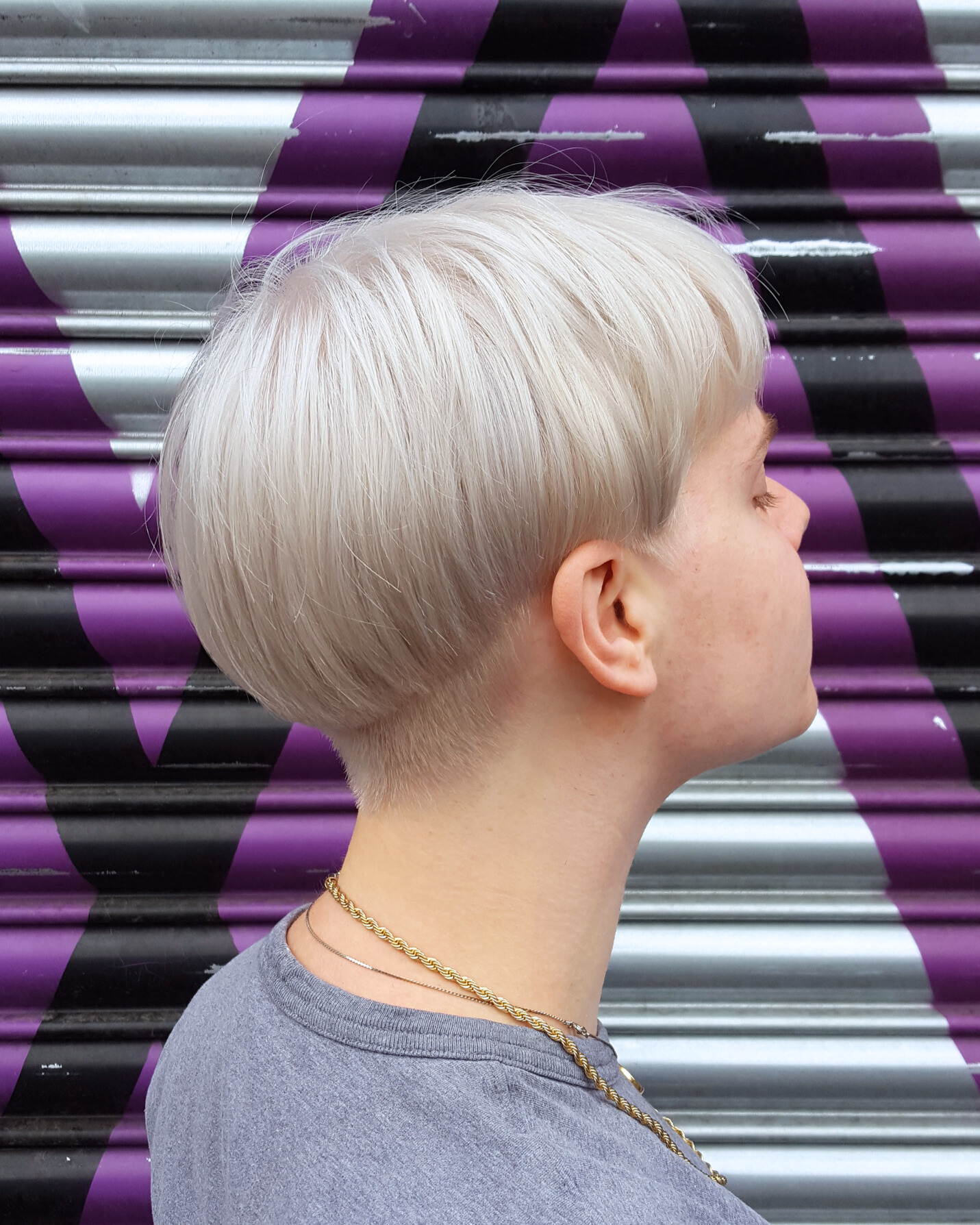 Side profile of a person with short, platinum blonde hair and a gold chain necklace, standing against a purple and black striped metal shutter.