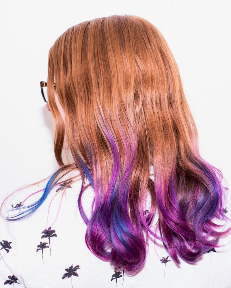 Person with long, wavy brown hair dyed purple and blue at the ends, wearing glasses and a white shirt with black palm tree prints.