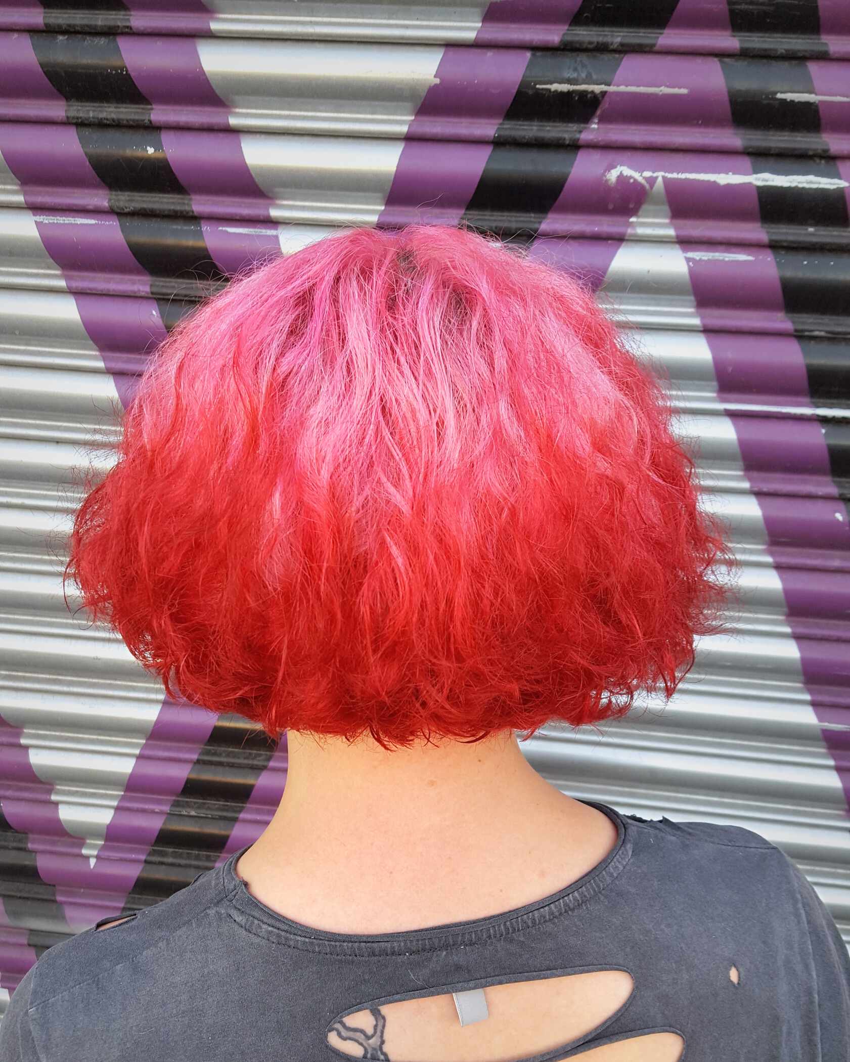 Person with short, wavy pink and red hair standing in front of a striped metal shutter with purple and black graffiti, wearing a black shirt with cut-out back.