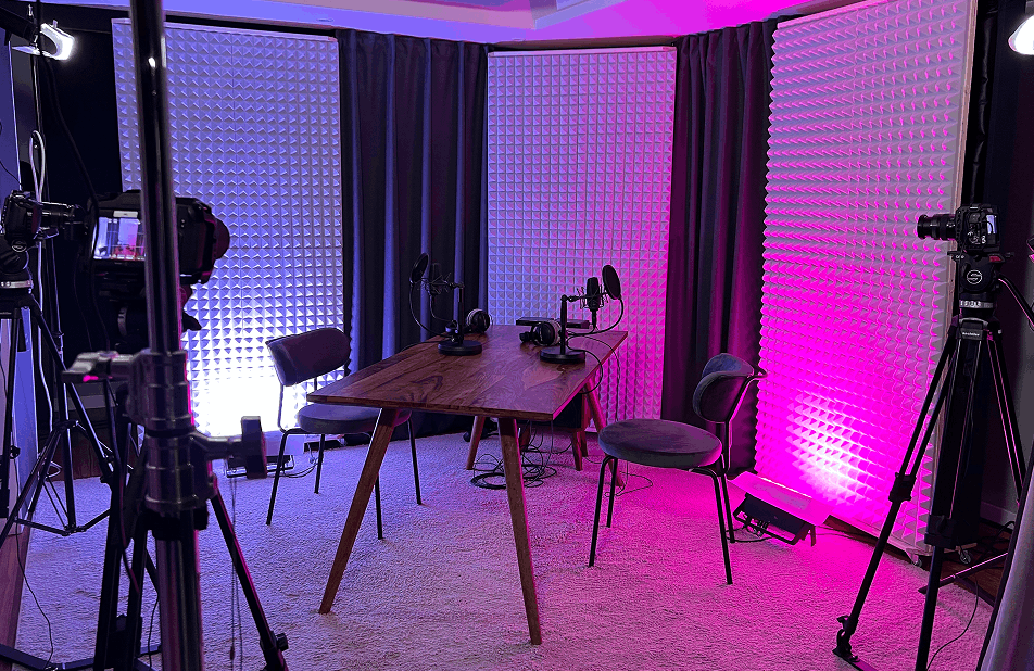 Podcast-Studio mit einem Holztisch, drei Mikrofonen mit Popschutz, drei Stühlen und Kameras auf Stativen, beleuchtet mit blauem und pinkem Licht.