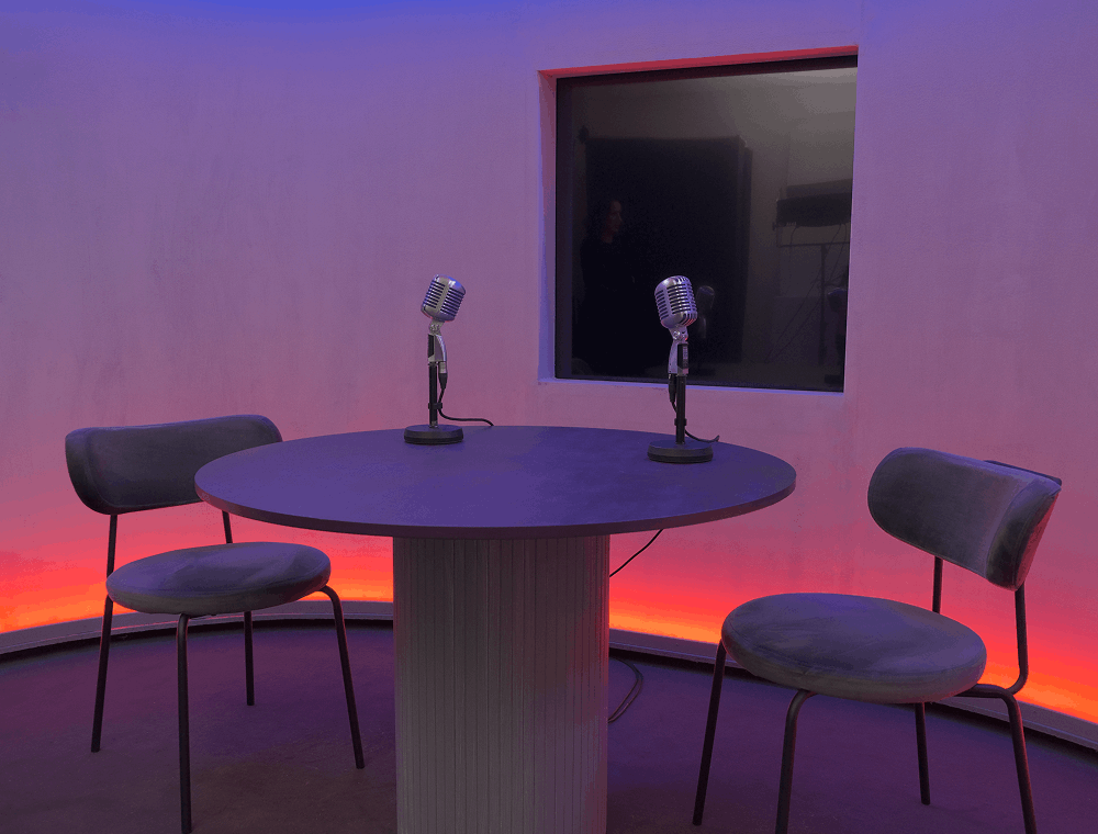 Runder Tisch mit zwei Mikrofonen und zwei Stühlen in einem Raum mit orange-blauer Lichtstimmung.