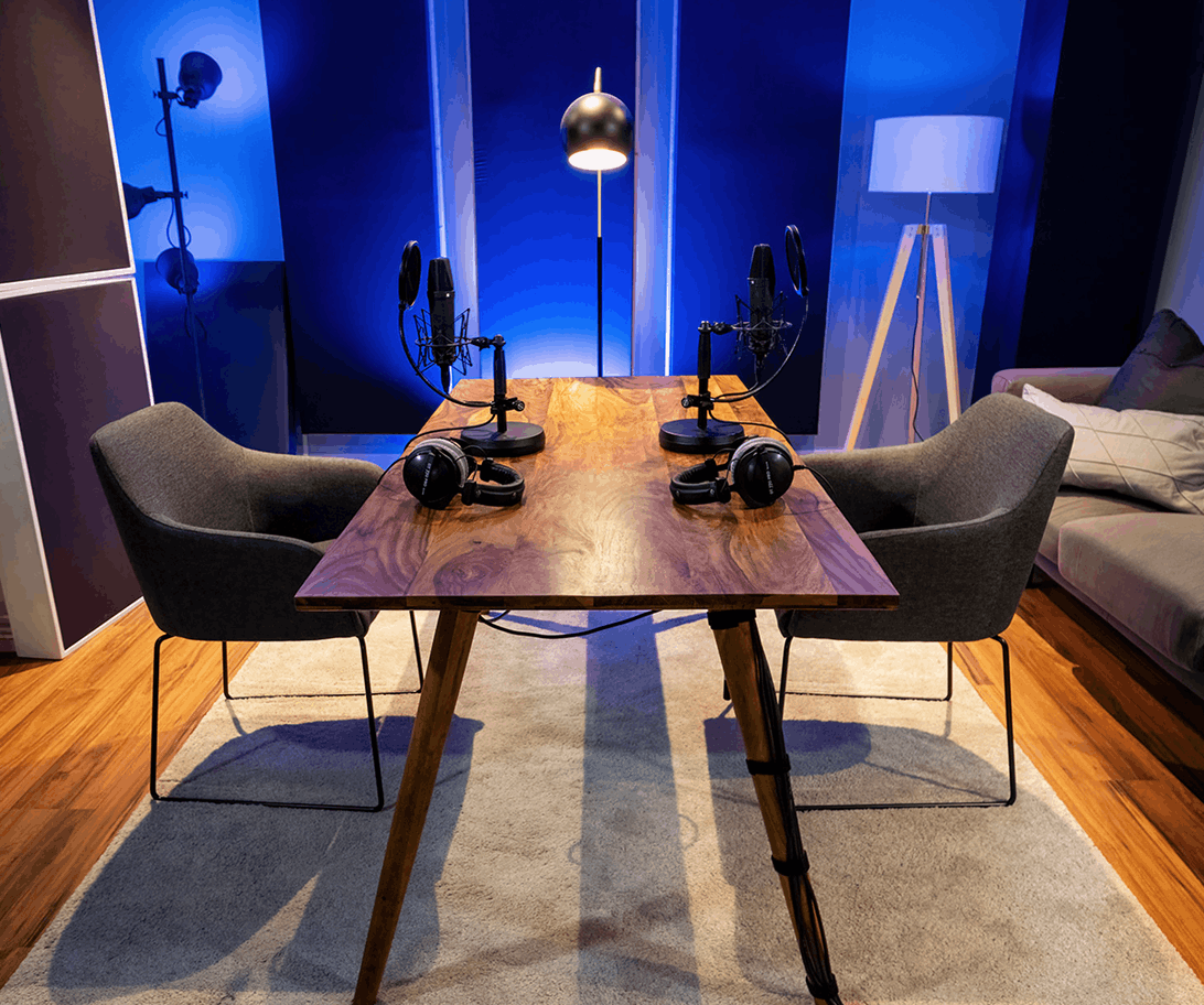 Podcast-Aufnahmeraum mit zwei Mikrofonen, Kopfhörern und Stühlen an einem Holztisch, blaue Akzentbeleuchtung im Hintergrund.