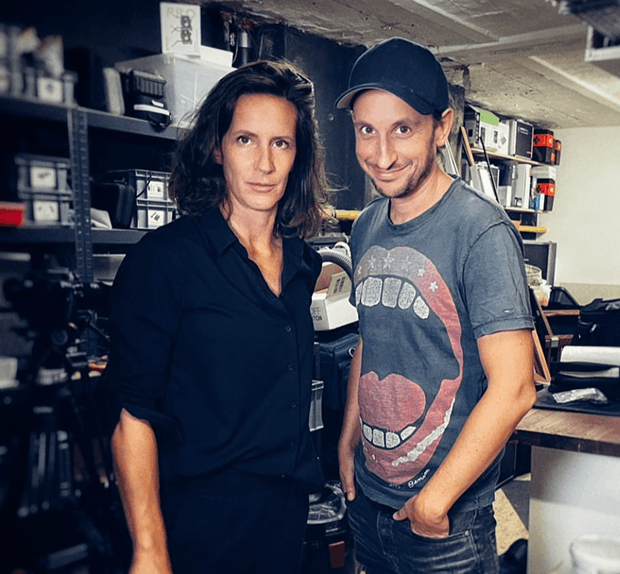 Su Holder und Chris Guse stehen in einem Raum mit Regalen voller Ausrüstung und Werkzeugen, eine Frau in schwarzem Hemd und ein Mann mit Kappe und T-Shirt mit offenem Mund-Motiv.
