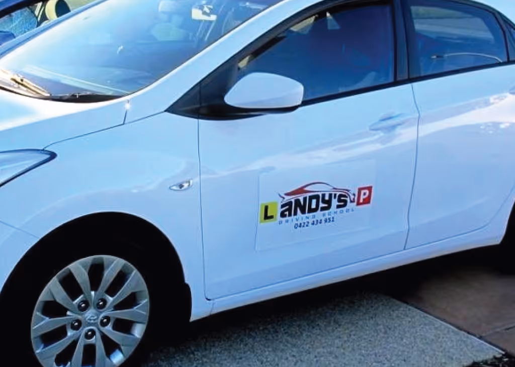 White car with Landy's Driving School logo on the front door parked on a driveway.