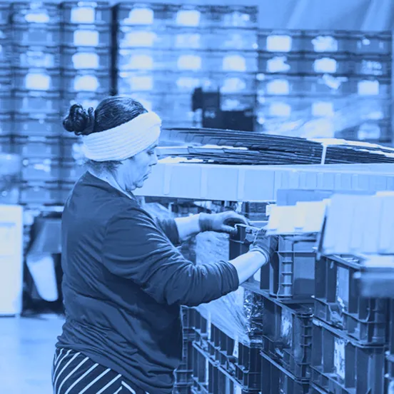 Worker in gloves loads flattened cardboard and packs items into red reusable plastic totes at a work table.