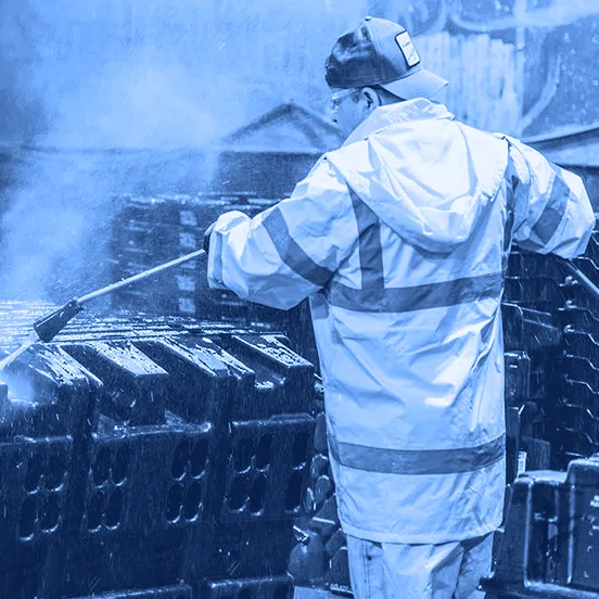 Worker in a high-visibility jacket uses a pressure washer to clean stacked plastic containers outdoors.