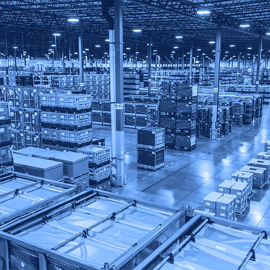 An overhead view of a large warehouse showing rows of stacked plastic totes, metal racks, pallets of boxed goods, and wide aisles under a high industrial ceiling.