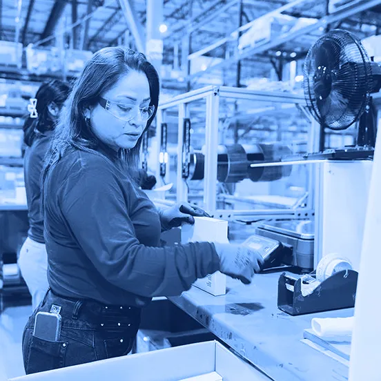 A worker wearing safety glasses and gloves scans a small box at a packing station inside a warehouse with shelving and equipment in the background.