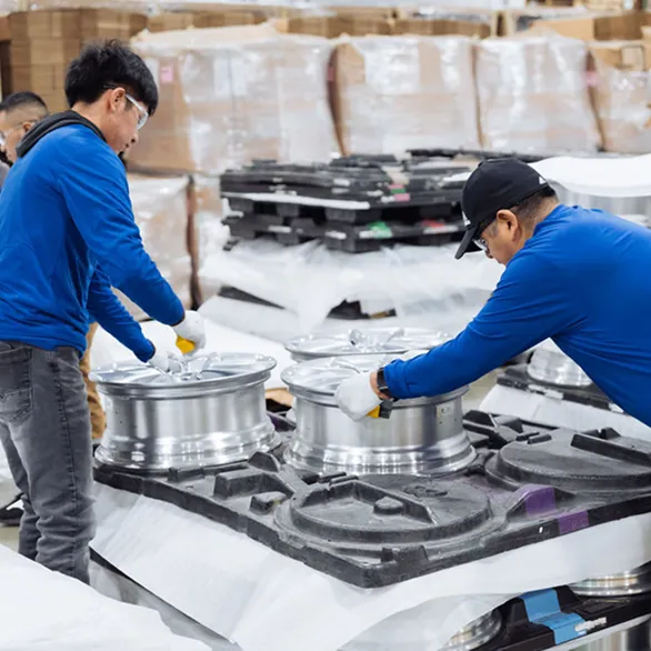 Two workers place silver wheel rims into molded trays on a pallet surrounded by stacked packaging materials.