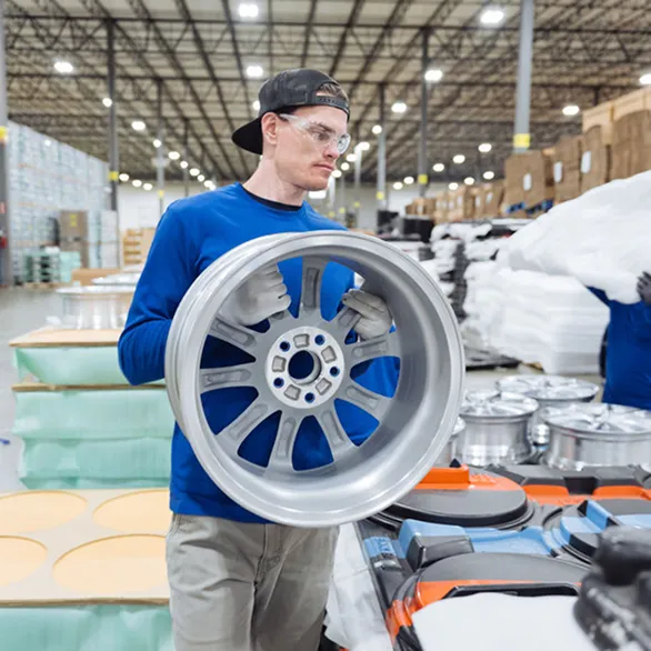 Worker in safety glasses holds a silver wheel rim while standing beside stacked trays of rims in a warehouse.