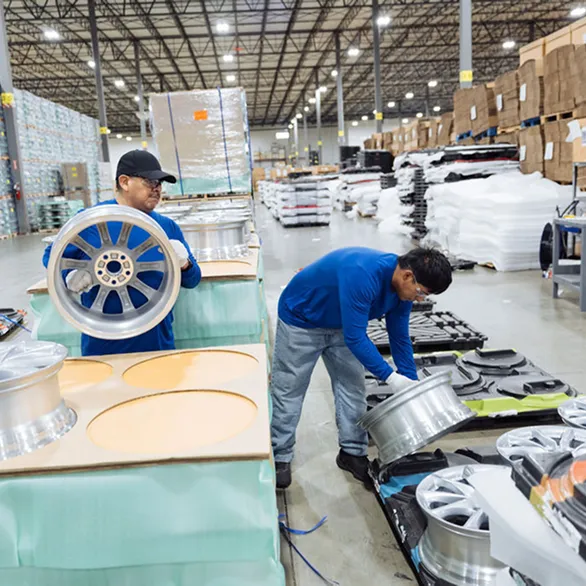 Two workers place silver wheel rims into a black plastic tray on a pallet in a warehouse aisle.