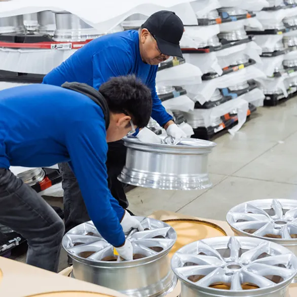 Worker in a blue shirt and safety glasses lifts a silver wheel rim from a pallet while another worker positions rims onto a tray.