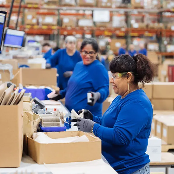 Worker wearing gloves places a white box into an open cardboard shipping box at a workstation.