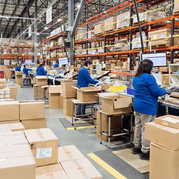 Worker wearing safety glasses places a small white box onto a scale next to a packaging machine.