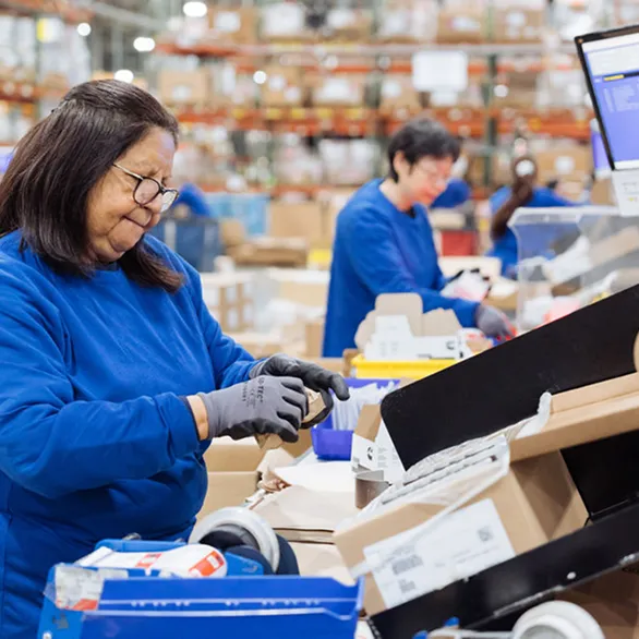 Worker wearing safety glasses operates a control panel next to a machine with spooled material and a small fan on the workstation.